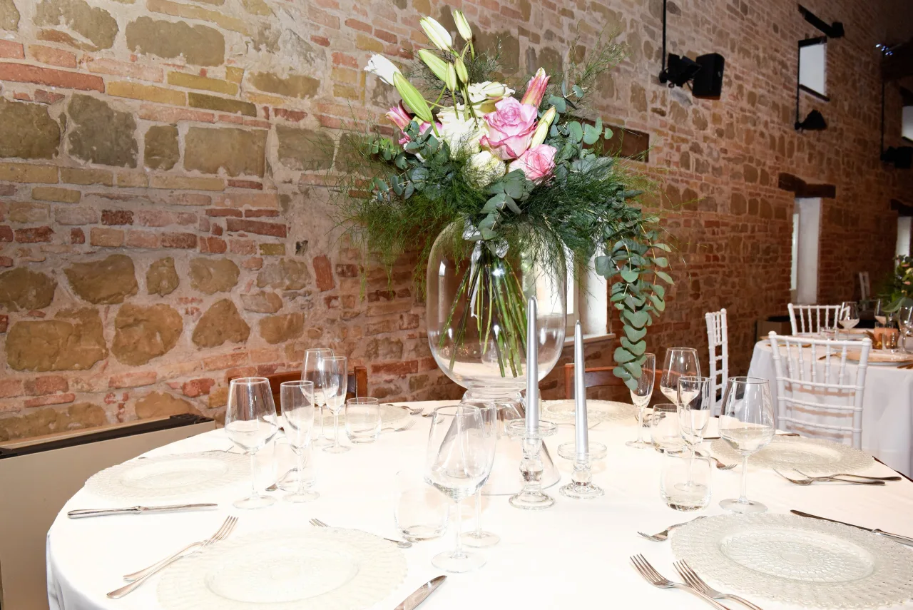 Tavolo rotondo apparecchiato in bianco con alto centrotavola di rose, gigli ed eucalipto e sedie Chiavari, contro parete in mattoni a vista di Villa Luzi