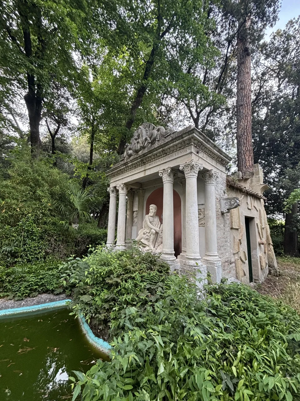 Il tempietto neoclassico del parco con statua seduta tra le colonne corinzie, immerso nella vegetazione accanto a una vasca d'acqua