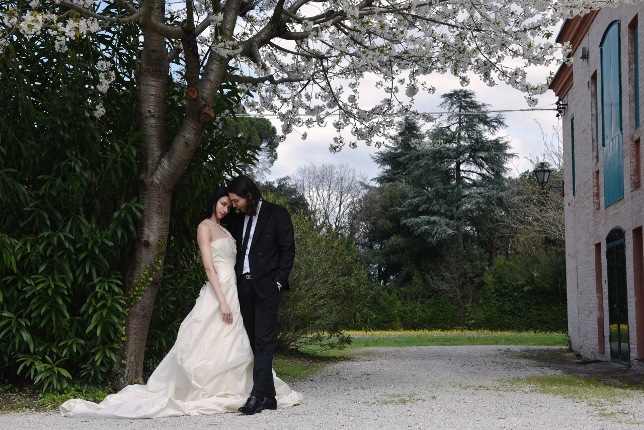 Sposi abbracciati sotto un ciliegio in fiore nel parco di Villa Luzi, con il corpo in mattoni della dimora e la campagna marchigiana sullo sfondo