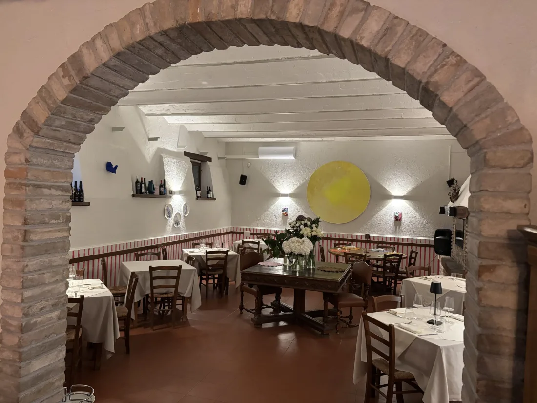 Sala da pranzo della dimora vista attraverso l'arco in mattoni a vista, con tavoli apparecchiati in bianco e disco dorato illuminato sulla parete avorio