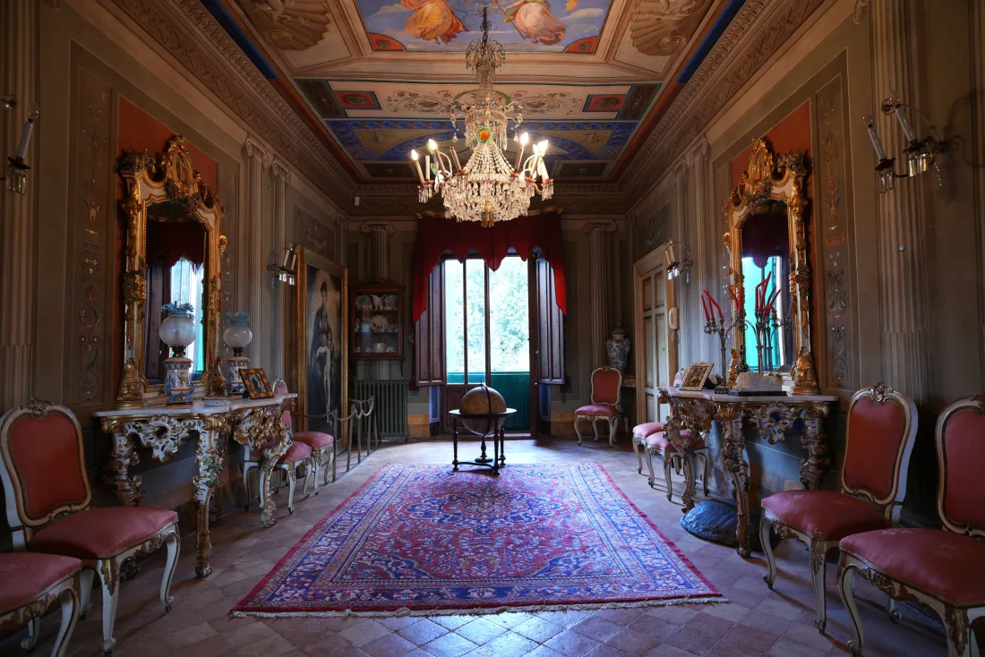 Salone della dimora con soffitto affrescato, lampadario di cristallo e sedute tappezzate disposte simmetricamente
