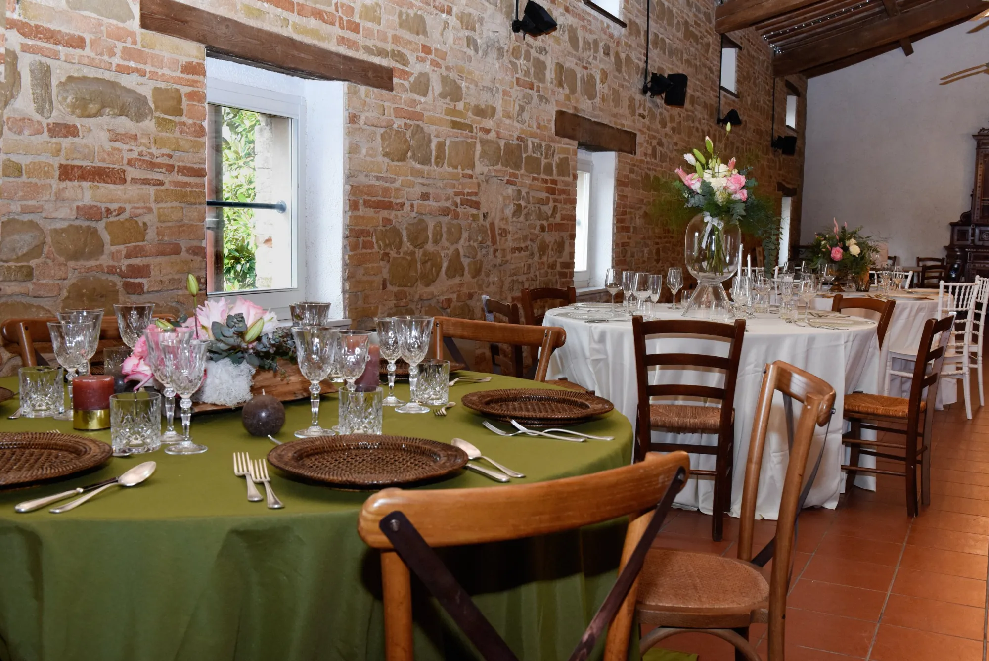 Sala da pranzo con pareti in pietra a vista allestita per un evento: tavoli rotondi apparecchiati con tovaglie verdi e avorio, calici di cristallo, piatti su sottopiatti in rattan e centrotavola floreali rosa